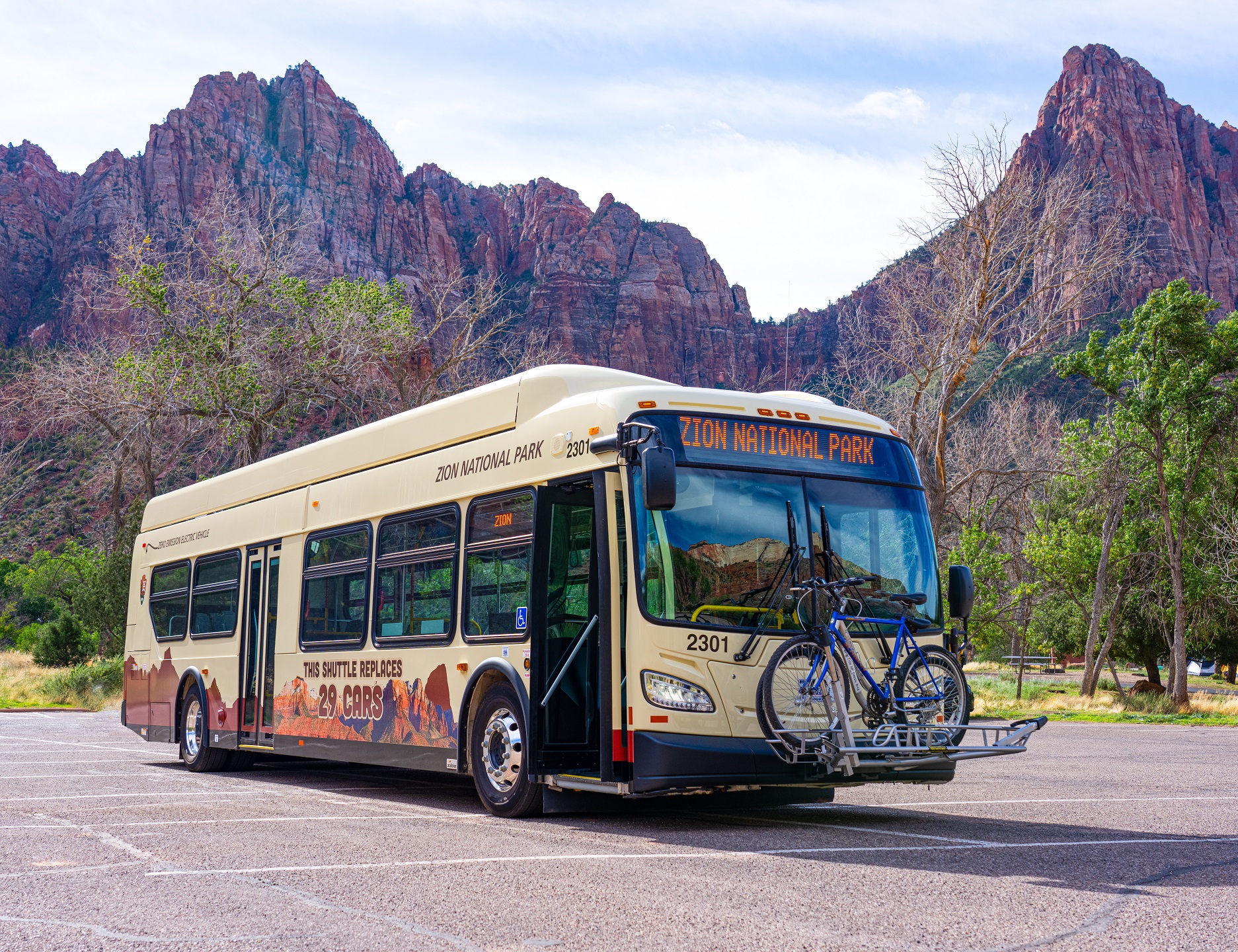 Picture of a full-size Zion National Park Bus in a parking lot with the mountains of the park in the background. The bus has a bike rack on the front and says Zion National Park on the front banner. On the side it says "This shuttle replaces 29 cars".